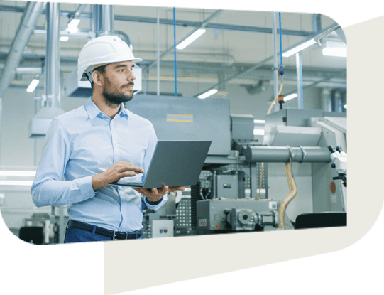 A professional man wearing a white hard hat and light blue shirt is standing in an industrial facility, holding a laptop