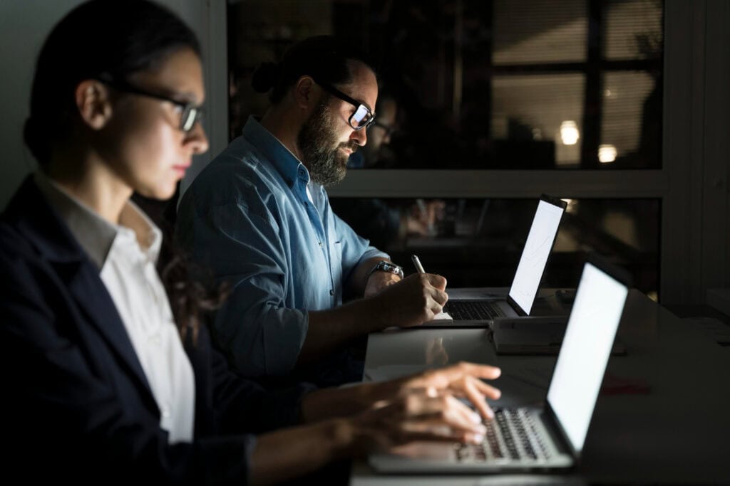 Two IT experts, one woman and one man, work in dark room lit only by their laptop screens