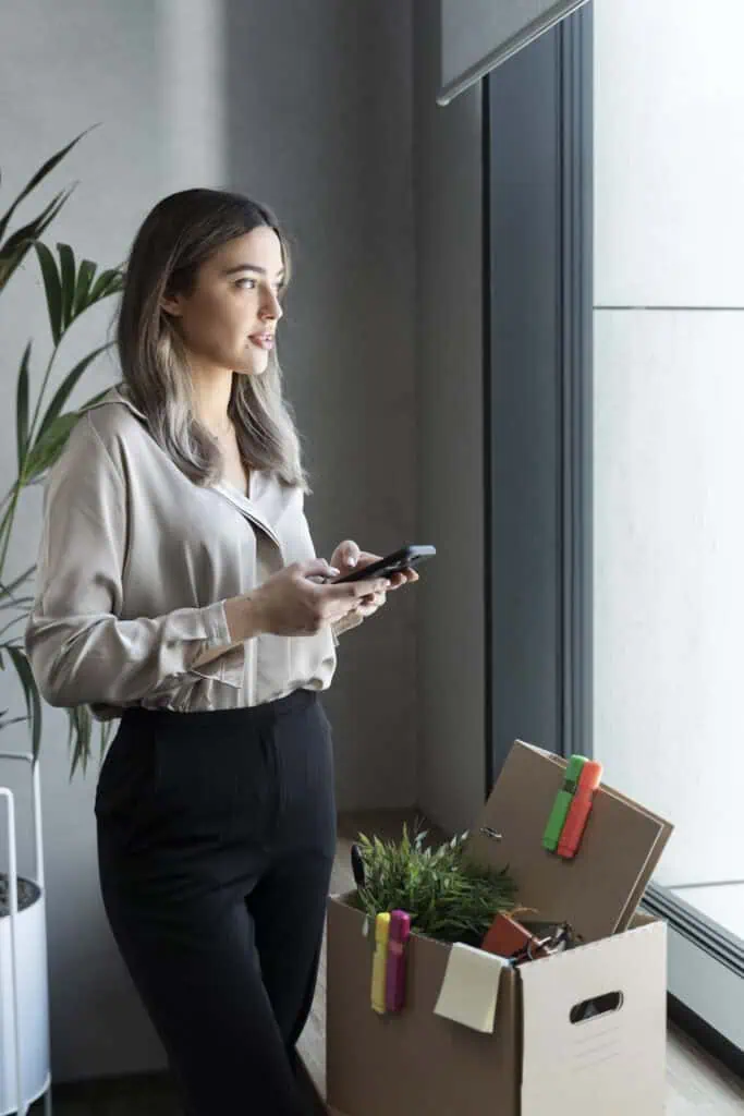 Relocation manager looking out of window with phone in her hands and box with her stuff sitting on the window ledge