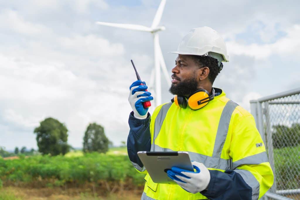 Man in a yellow safety vest is holding a walkie talkie and a tablet. He is standing in a field with a wind turbine in the backround