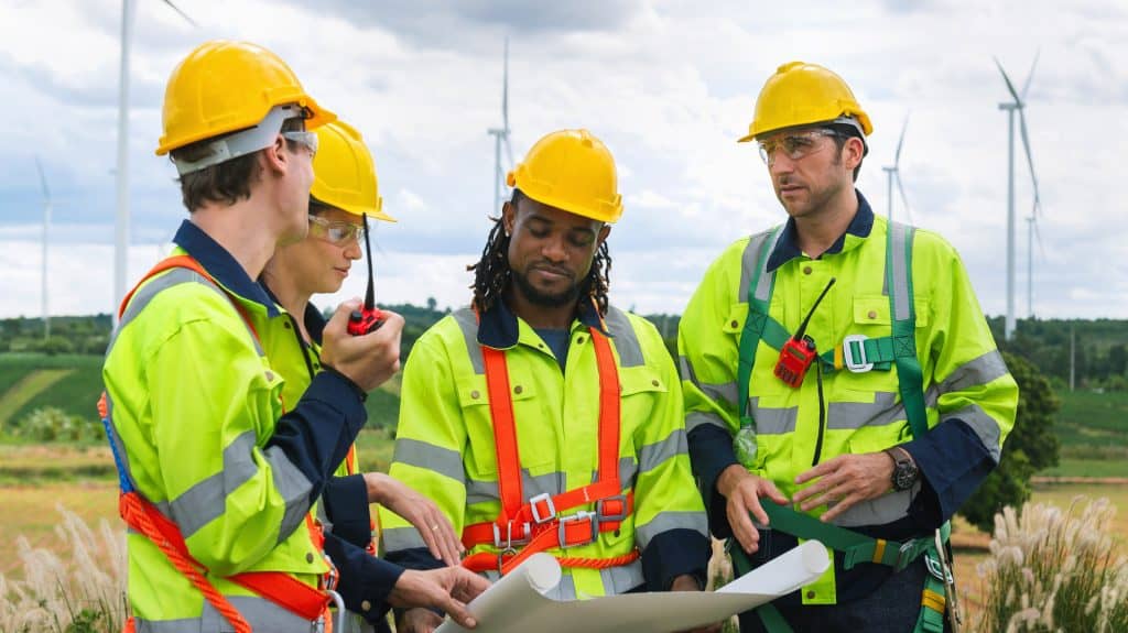 Group of construction workers wearing yellow vests and hard hats are standing around piece of paper with wind tourbines in the backroud